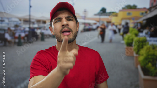Canvastavla Young hispanic man pointing finger toward viewer in red shirt and cap on bustling city street; accusation confrontation