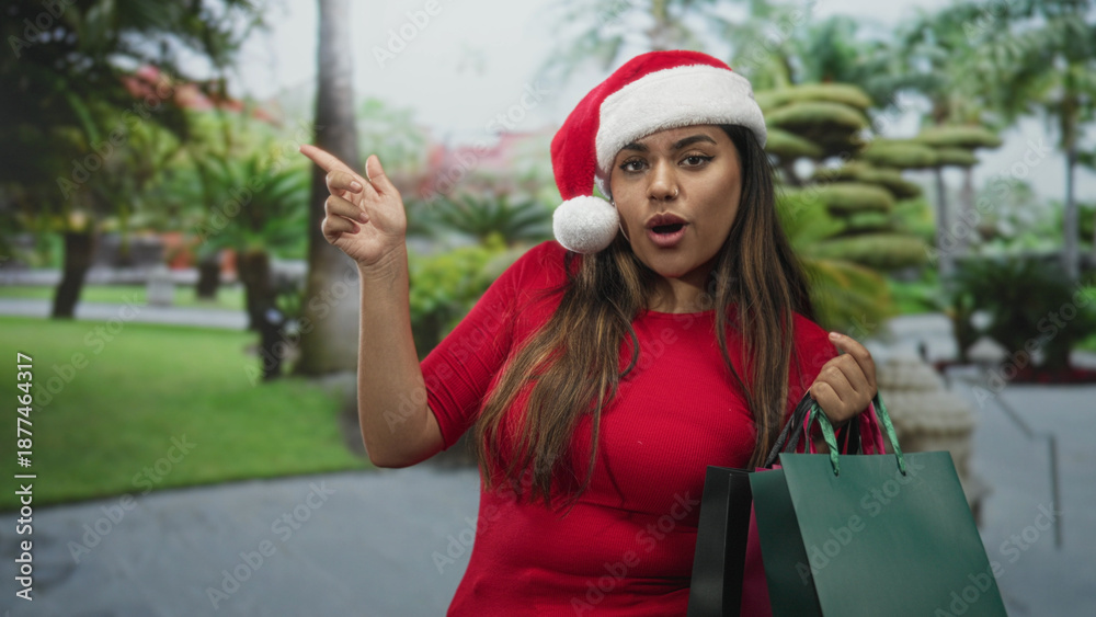 Obraz premium Woman holding shopping bags points finger to left on street while wearing santa hat and smiling; holiday cheer.