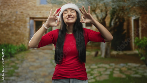 Young woman in santa hat makes ok hand signs with both hands and smiles, standing on a cobblestone path before a stone building, casual red shirt and long black hair; joyful celebration.