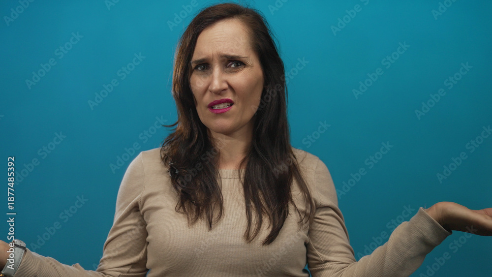 Obraz premium Woman with confused expression against blue background wearing beige sweater with long brown hair raising hands in questioning gesture capturing unsure emotion.