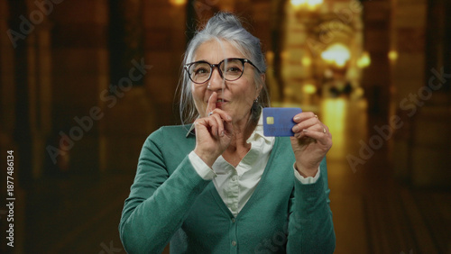 Senior woman with grey hair holding credit card in church while gesturing silence, wearing glasses and green sweater, indoor scene providing contrast and mystique.