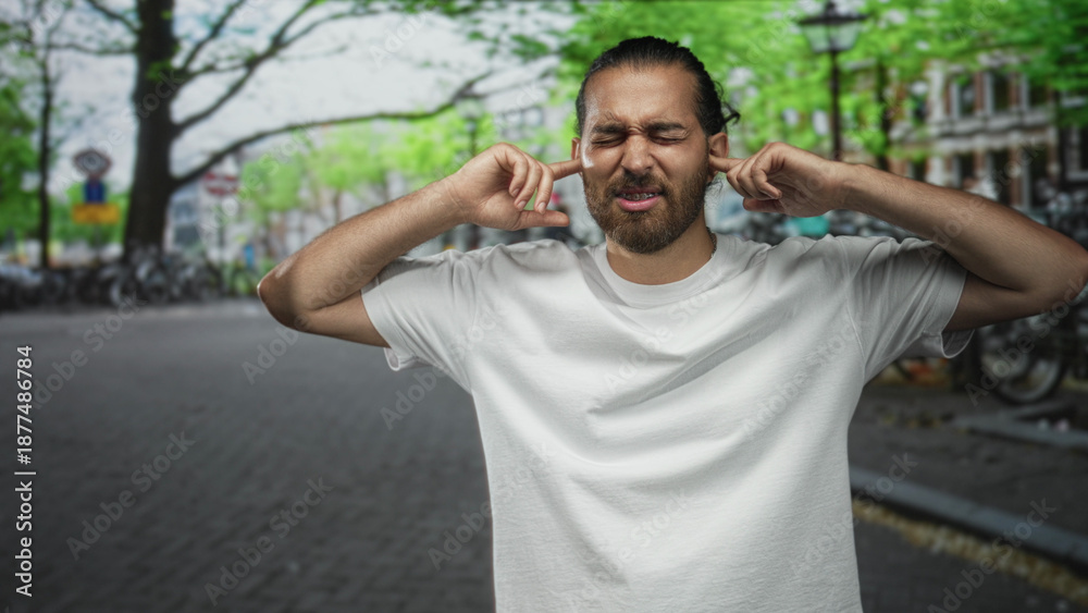 Naklejka premium Man with beard pressing fingers into ears on a crowded city street, eyes closed and grimacing; frustration overload.