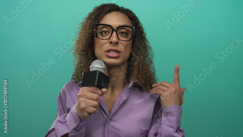 Bilde på lerret Woman with curly hair wearing glasses and a purple shirt holds microphone and points finger in teal studio; confidence