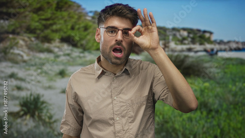 Man holds hand in ok sign near eye at beach under bright daylight by shore; serenity approval confidence.