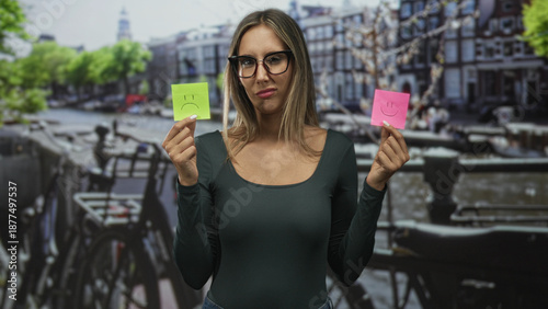 Tableau sur toile Woman holding two sticky notes with smile and frown faces on a canal street in amsterdam, wearing glasses and showing ambivalent expression; indecision