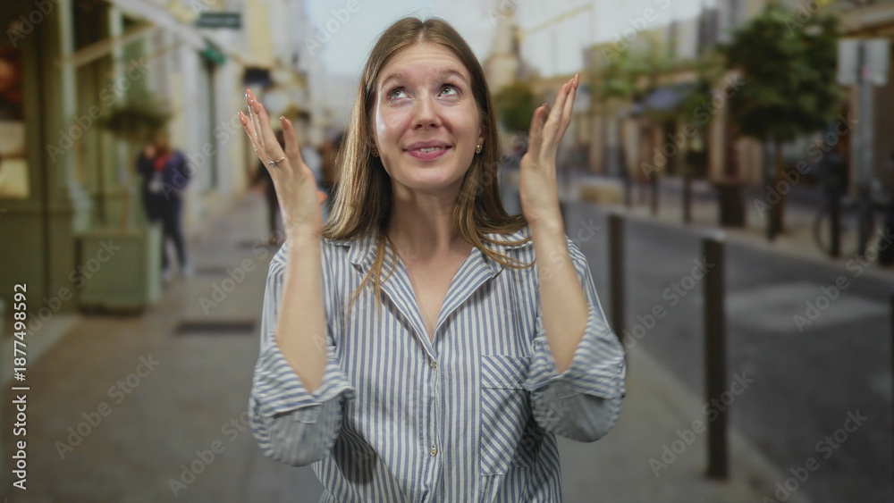 Fototapeta premium Woman with clasped hands smiling on street wearing striped shirt by storefronts and lampposts on sidewalk under city buildings in daylight; happiness.