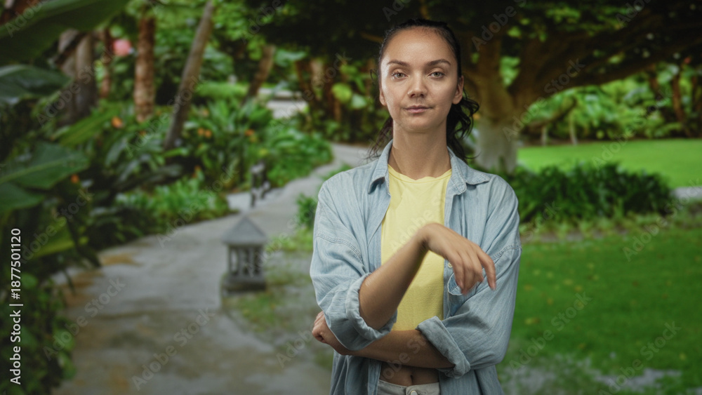 Fototapeta premium Woman standing with arms crossed showing bare forearms, wearing yellow shirt and denim overshirt on a garden path amid lush trees in forest; calm reflection.