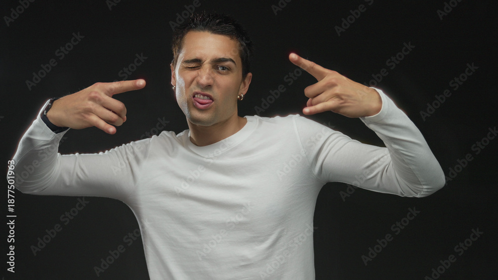 Naklejka premium Young hispanic man in white shirt shows rock horns hands gesture in studio; playful metal music spirit.