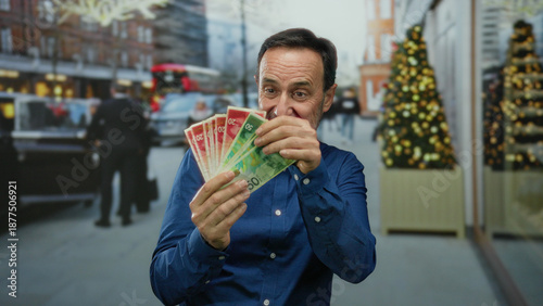 Photos Man holding israeli shekel banknotes, standing in an urban city street with festive decorations, expressing happiness outdoors