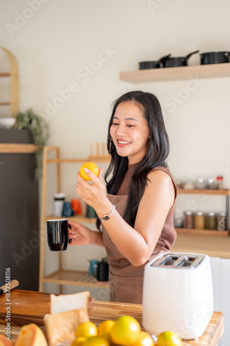 Pretty asian woman holding coffee mug as looking at orange standing aside toaster on kitchen counter