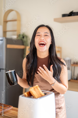 Laughing asian woman holding coffee mug and standing aside bread toaster on kitchen cooking counter.