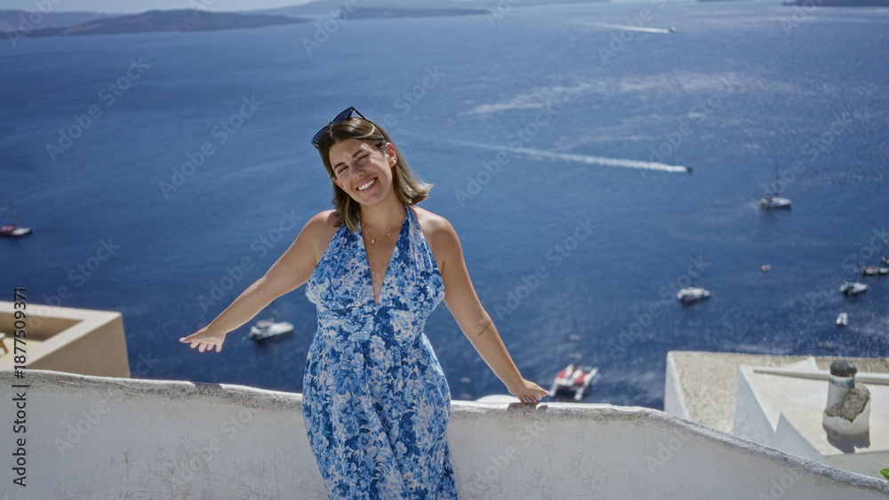 Fototapeta premium Woman smiling with arms out on building terrace overlooking sea and boats in santorini town; vacation relaxation joy.