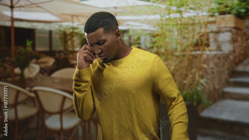 Valokuva Young black man holding phone to ear with tense frown in outdoor cafe patio wearing yellow sweater and leaning by stone steps; frustration urgency