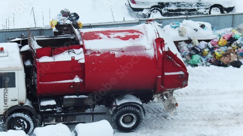 Waste collection vehicle processing garbage bags in winter environment, heavy snow blankets street and surroundings, difficult conditions for sanitation service, accumulation of trash bags near site