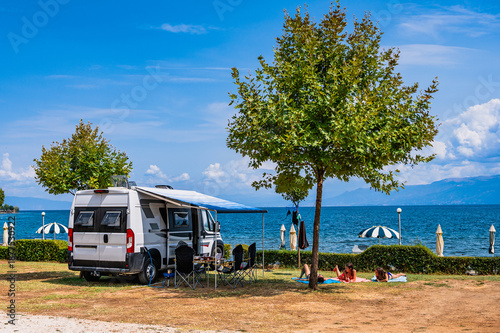 A scenic summer day at a Lake Ohrid campsite in Udenisht, Albania, featuring a camper van with an awning and a family sunbathing on the grass under a tree with clear blue waters in the background.