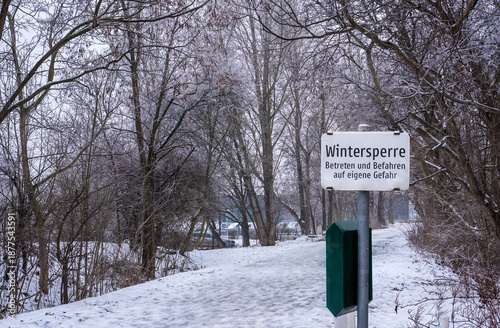 Canvas-taulu A snow-covered path in the inner city area near a parking lot, that has not been cleared of snow with a sign at the beginning that says in German: Winter closure, enter and drive on at your own risk
