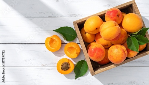 Overhead shot of ripe apricots, some halved, in a wooden box on a white wooden surface
