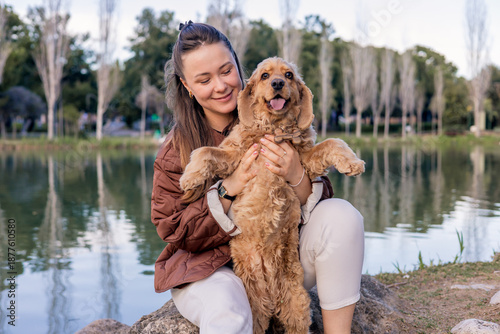 A young Caucasian woman with long brown hair holds a happy golden cocker spaniel by a serene lake surrounded by trees.
