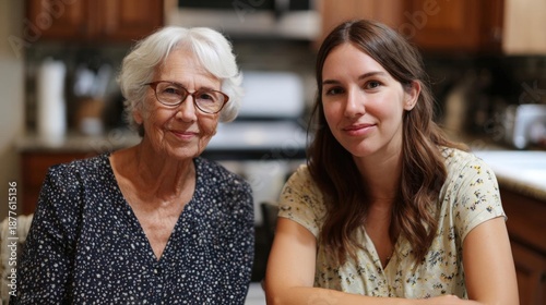 Elderly woman and a young woman sitting in a kitchen. the elderly woman is on the left side of the image, wearing a black dress with white polka dots and glasses.