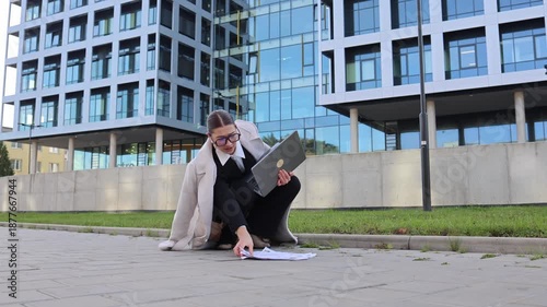 Young woman running late for work kneels to gather scattered papers while holding laptop rushed stressed moment near office building city morning pressure