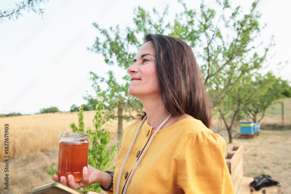 Fototapeta premium Side view of mature woman holding jar of honey in natural farm landscape with trees and field