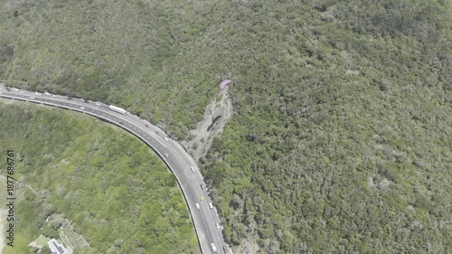 Drone lowers as camera pans up over coastal highway with paragliders above on a sunny day on the Garden Route in Wilderness, South Africa