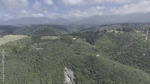 Drone flies backwards from paragliders over forested hll next to coastal highway on a sunny day on the Garden Route in Wilderness, South Africa