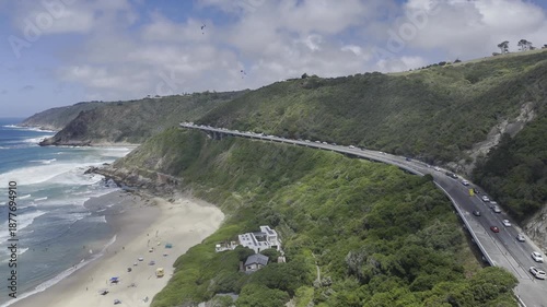 Drone lowers in epic shot of Wilderness Beach with paragliders on a sunny day on the Garden Route in Wilderness, South Africa