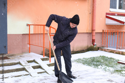 A middle-aged Caucasian man with short hair shovels snow outside a building. He wears a black beanie and a dark jacket. Snow covers the ground.
