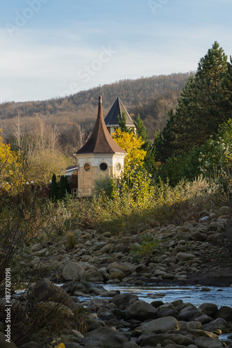 Country house is located on banks of mountain river. Octagonal tower with pointed roof and round windows, partially hidden by lush foliage, rises above autumn trees and is surrounded by cliffs