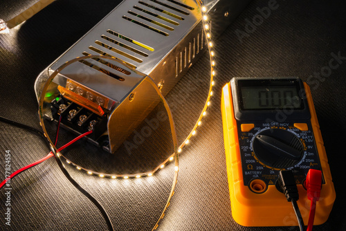 Photos a metal power supply unit connected to a coiled, illuminated LED strip emitting warm light, positioned next to a yellow digital multimeter displaying a zero reading on a dark textured surface