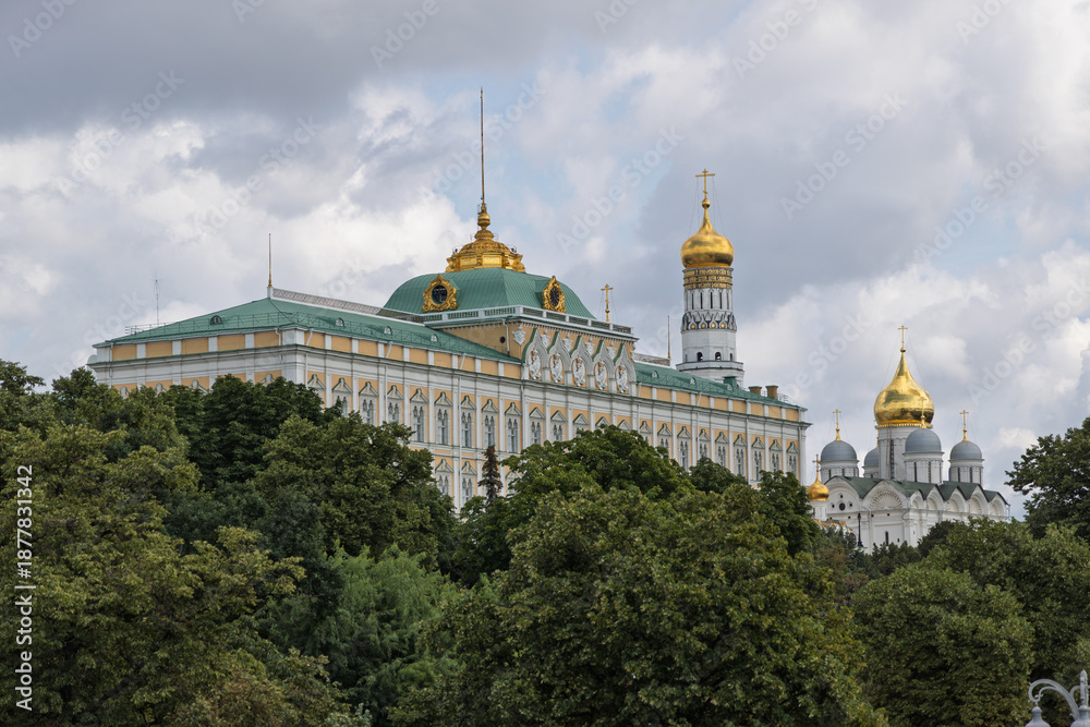 Fototapeta premium Grand Kremlin Palace and golden-domed cathedral complex in Moscow, framed by lush green trees under cloudy sky
