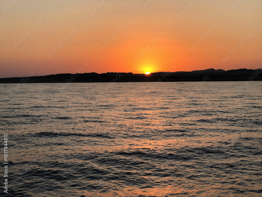 Naklejka premium Vibrant sunset over calm water with distant hills and orange sky