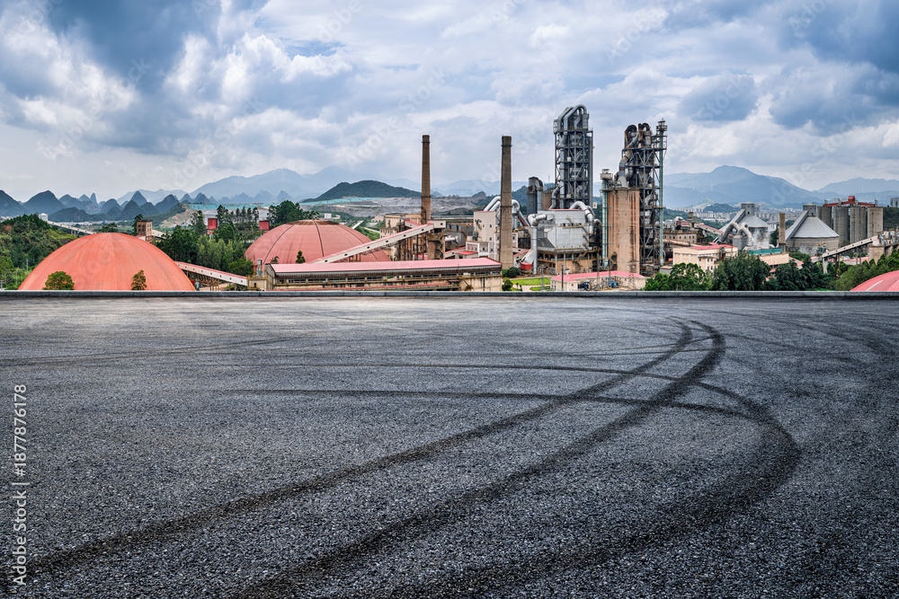 Obraz premium Industrial cement factory plant standing in a mountain landscape with an asphalt road and tire marks in the foreground.