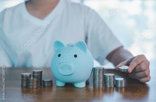 Fotografia Child saving money with blue piggy bank and stacked coins on table, symbolizing financial education, budgeting habits, smart savings, allowance management, and personal finance planning