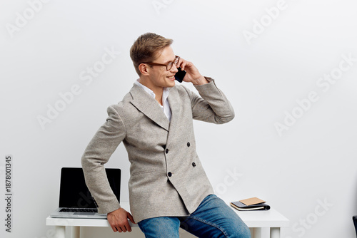 Friendly smiling young man wearing glasses and a stylish gray blazer sitting on a desk in a modern office, talking on mobile phone, cheerful mood, professional looking, casual yet elegant style