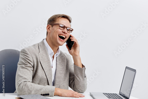 Professional middle-aged man in smart casual outfit laughing happily while talking on mobile phone, sitting at office desk with laptop, cheerful expression, isolated on plain white background