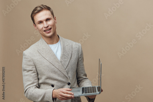 Smiling young man holding a laptop computer in a studio setting, wearing a beige blazer and light blue shirt, isolated on a plain beige background, casual professional appearance, technology concept