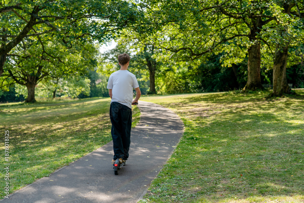 Fototapeta premium Boy rides electric scooter along path in green park during sunny day