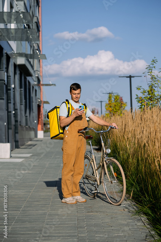 Foto Young male bike courier in brown overalls and yellow delivery bag stands with bi