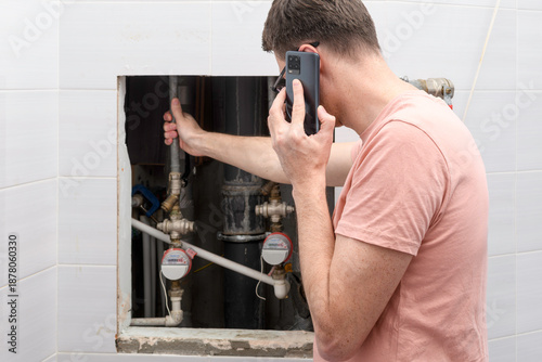 Fotografi Man calls emergency plumber beside open utility hatch, hand on pipe near water meters, reports leak in apartment bathroom