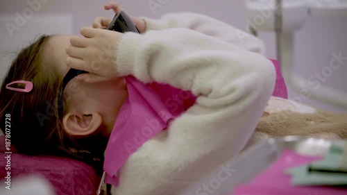 A young girl lies back during a dental visit, her thoughtful expression and the bright pink drape creating a striking scene.