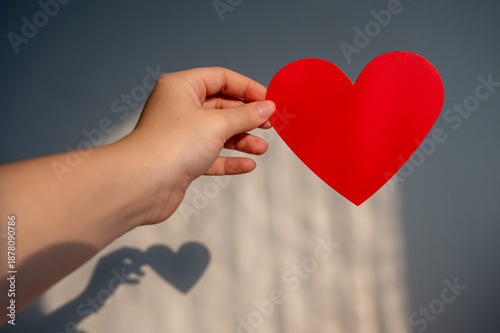 Hand holding a red paper heart against a grey wall with a beautiful shadow. Romantic Valentine's Day or charity concept