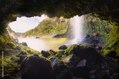 A moss covered cave opens toward a bright lagoon and cascading Lagoa do Vento waterfall on Madeira Island, Portugal, with wet rocks framing the scene in soft, diffused light