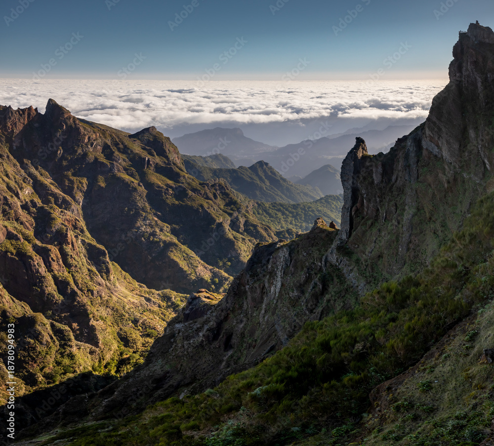 Fototapeta premium Warm morning light highlights rugged green mountains on Madeira Island, Portugal, as they rise above a vast sea of clouds, seen from Pedra Rija viewpoint under a clear blue sky