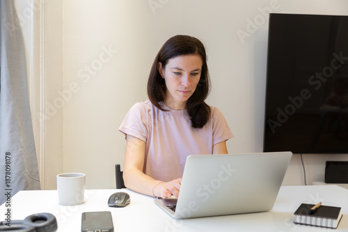 Young woman with long dark hair is working on a laptop at a bright desk, with a coffee cup and notebook nearby, showcasing a productive home office environment