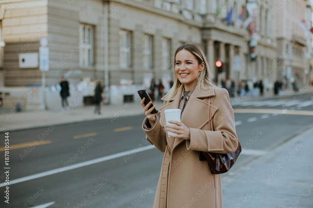 Fototapeta premium Woman holding coffee and phone while standing on city street with buildings