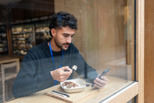 Quadro em tela Young man having lunch, eating food from a disposable container while browsing c