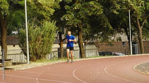 A man in sportswear jogs along a stadium track surrounded by trees and stops as sudden leg pain forces him to slow down. He begins to limp and heads toward a nearby bench to sit and rest