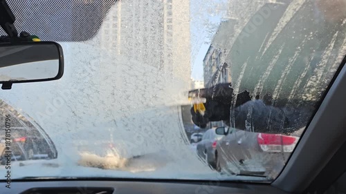 View from the interior of an automobile to a man in warm clothes cleaning the windshield of a car from snow, using an ice scraper. Winter car maintenance. 